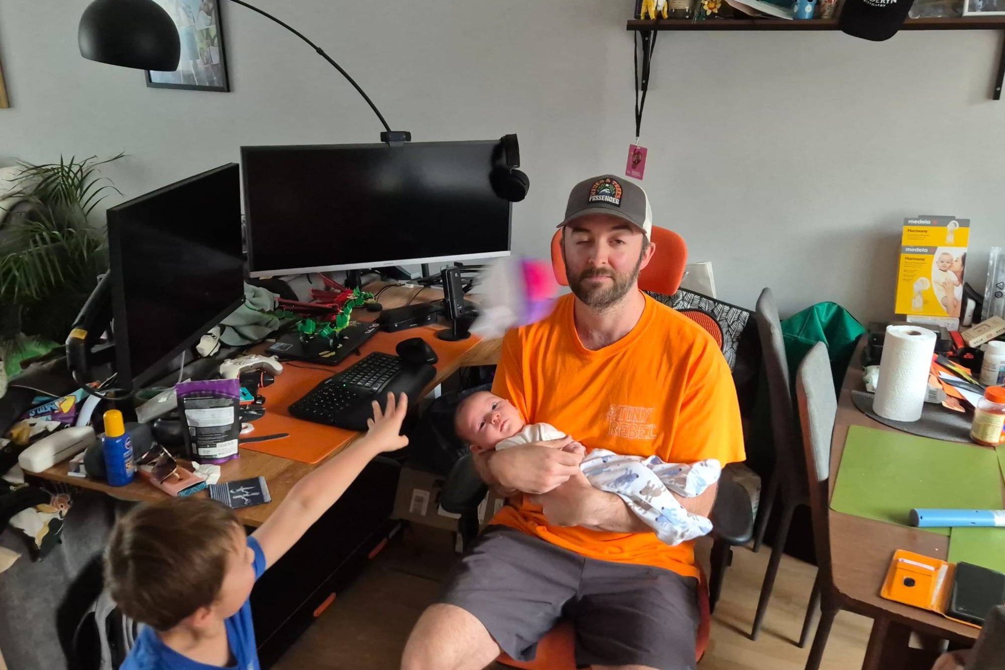 Dave at his desk in an orange T-shirt and cap, holding his newborn, while his four-year-old throws a toy Master Ball at his head. The ball is a purple blur. Behind them: two monitors, a Lego dragon, and a desk and table covered in family clutter.